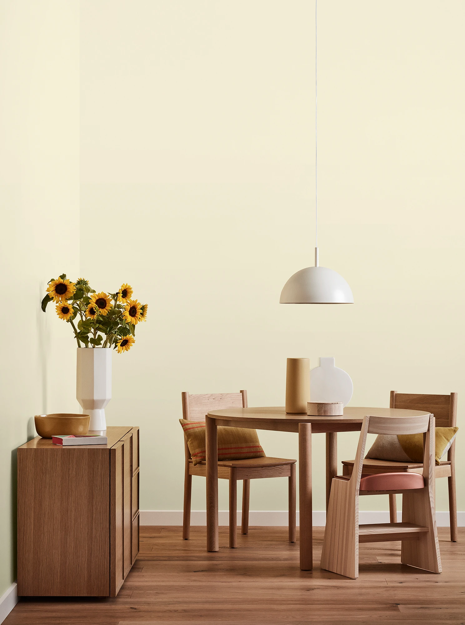 Round wooden table, three chairs and console with vase of sunflowers and white pendant light in neutral room.