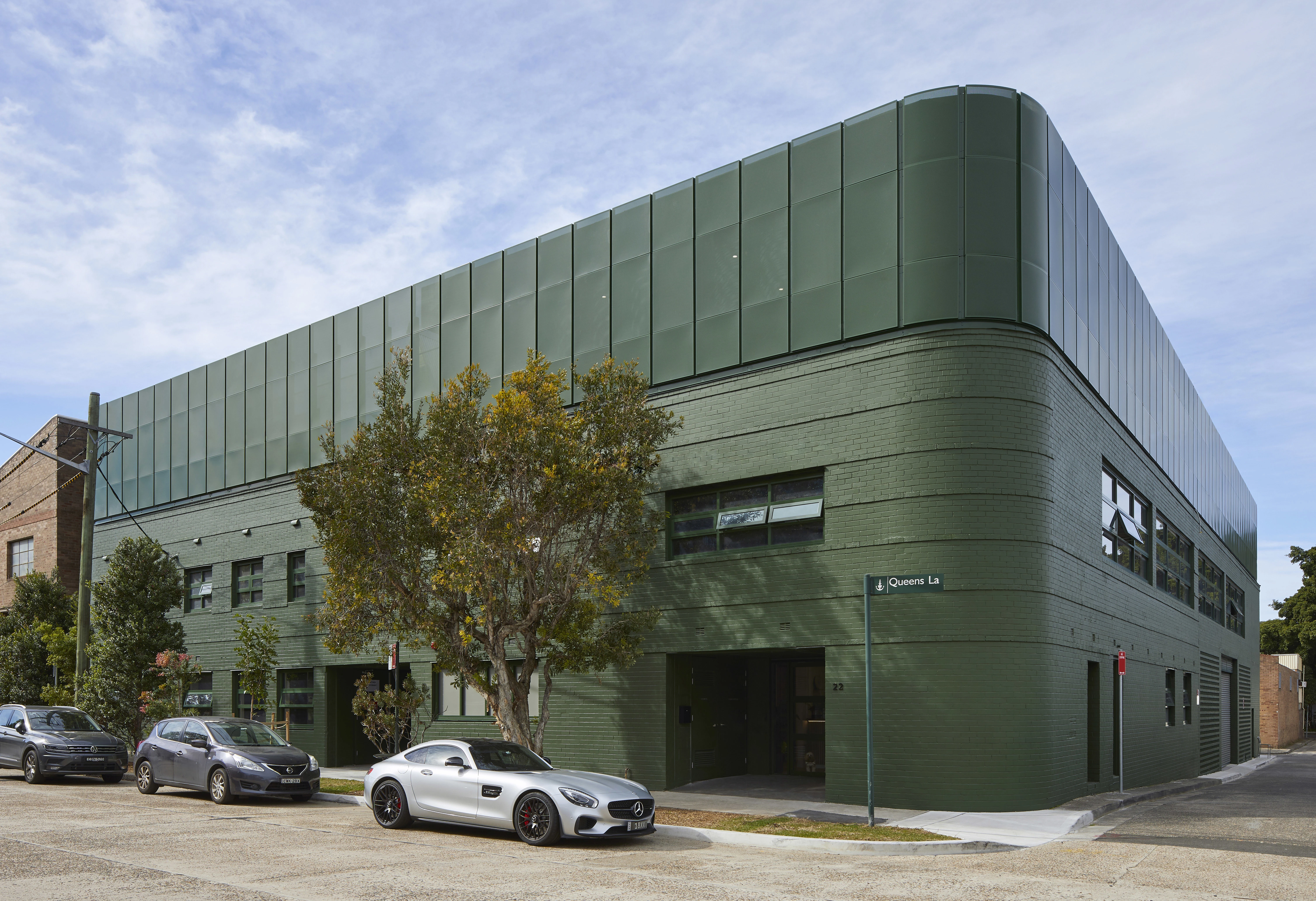 Green industrial building featuring luxury cars parked in front of it. 