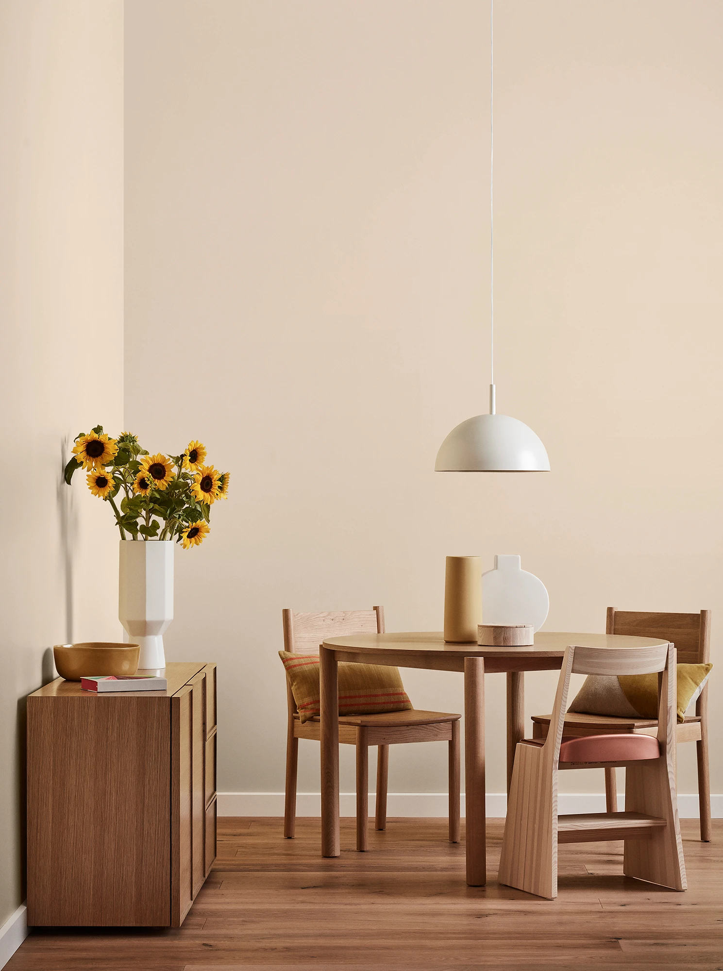 Round wooden table, three chairs and console with vase of sunflowers and white pendant light in neutral room.