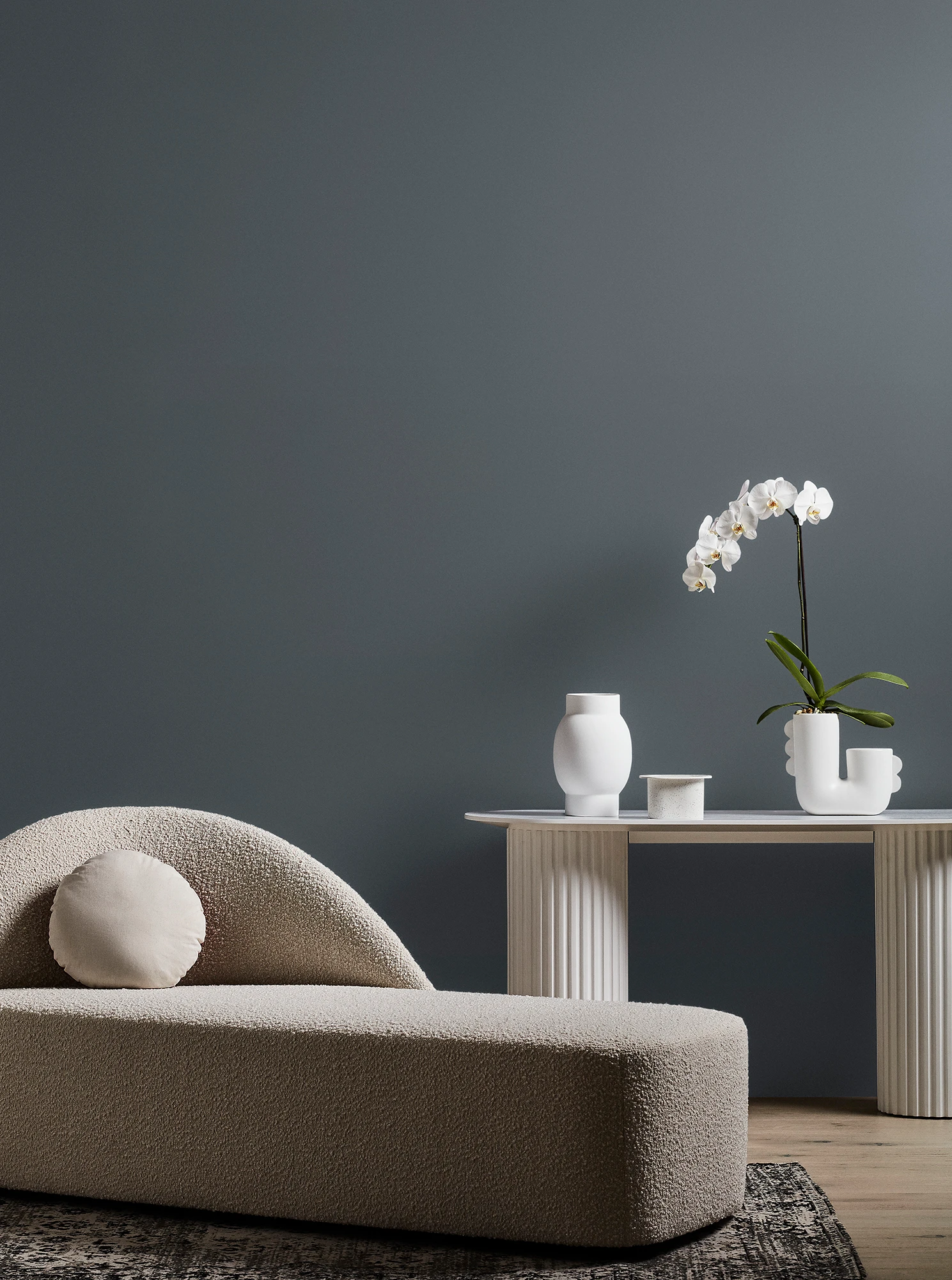 Neutral-coloured modern chaise longue and white table with orchid plant and vase in grey living room.