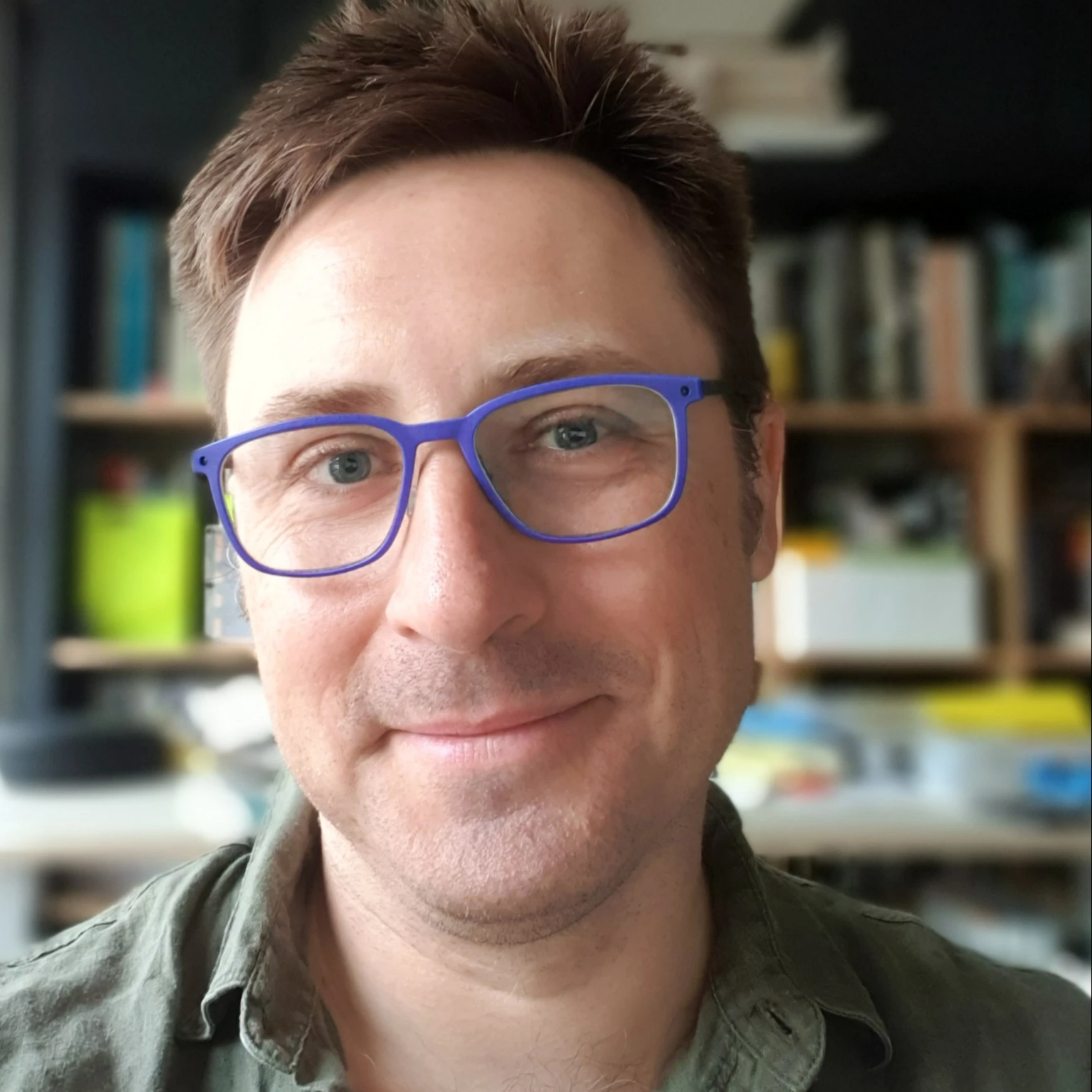 Adam Pustola headshot. Man with short brown hair and blue glasses smiling.