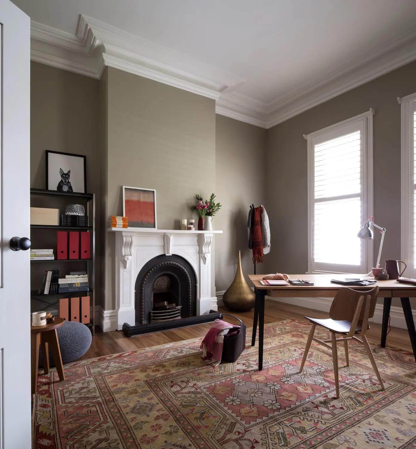 Study with green walls and fireplace. There's a patterned mat covering most of the wooden floor and a wooden table and chair with a white lamp on it.