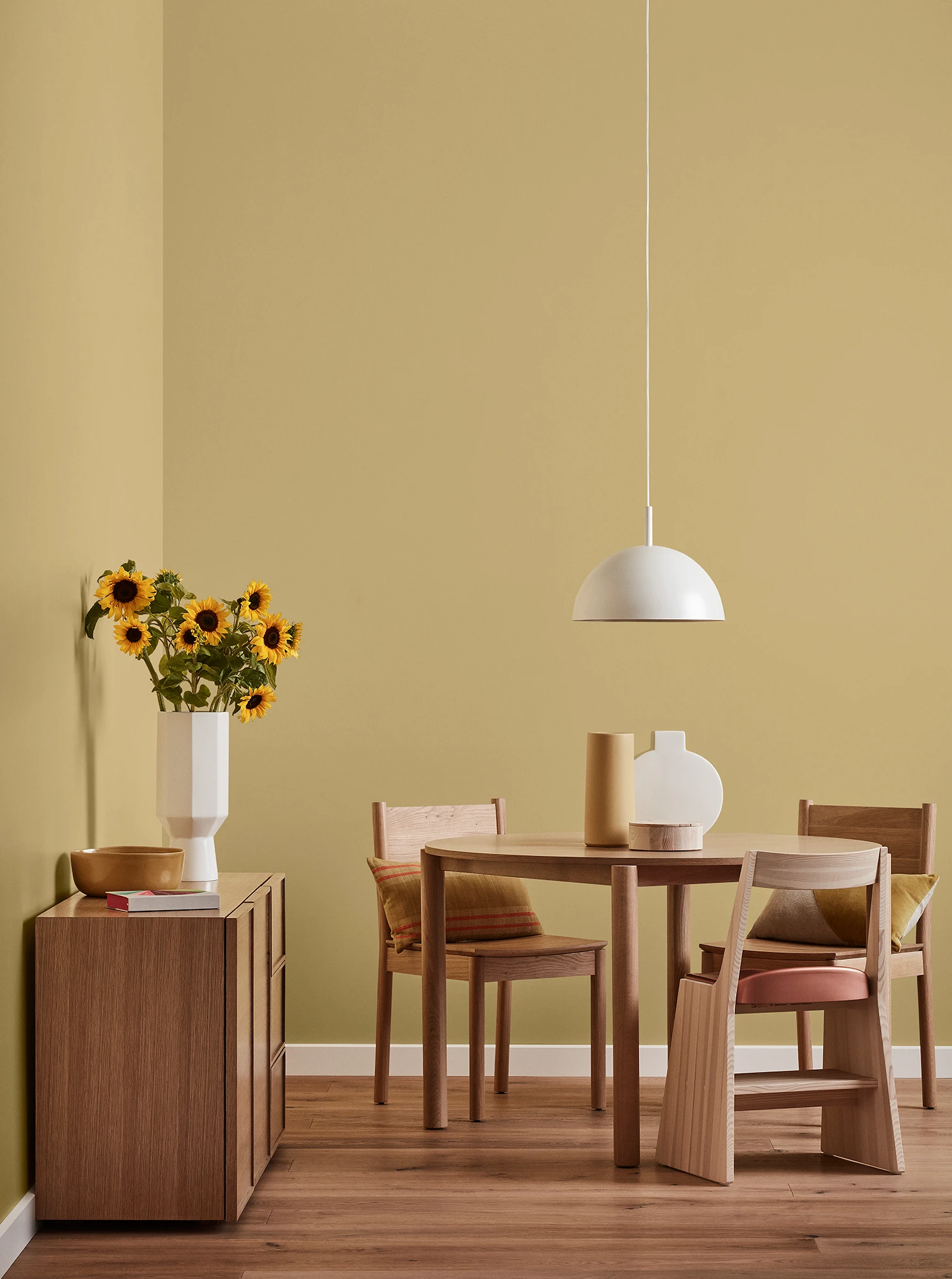 Round wooden table, three chairs and console with vase of sunflowers and white pendant light in yellow-brown room.