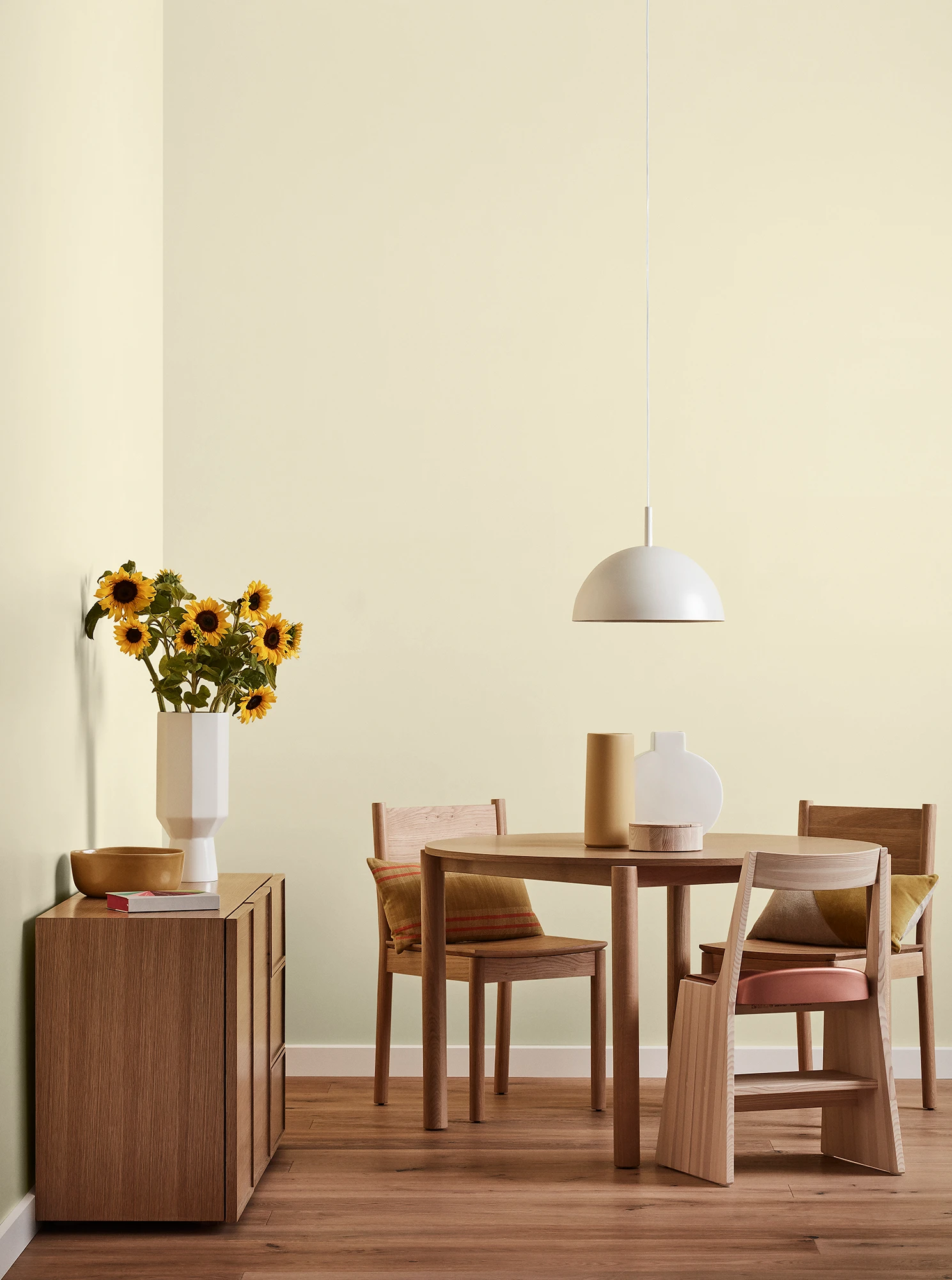 Round wooden table, three chairs and console with vase of sunflowers and white pendant light in neutral room.