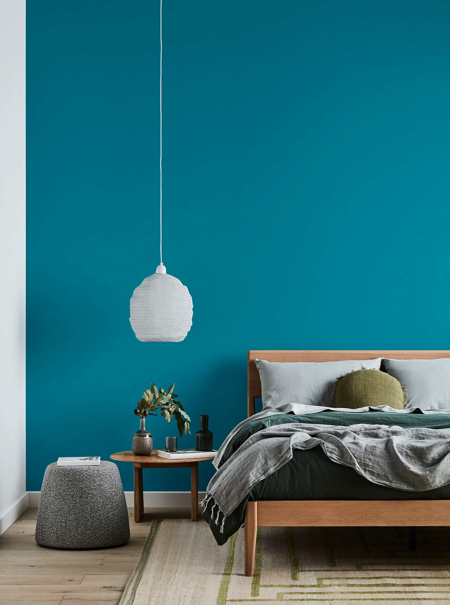 Green bedroom with timber bed and side table, white pendant light and grey linens.