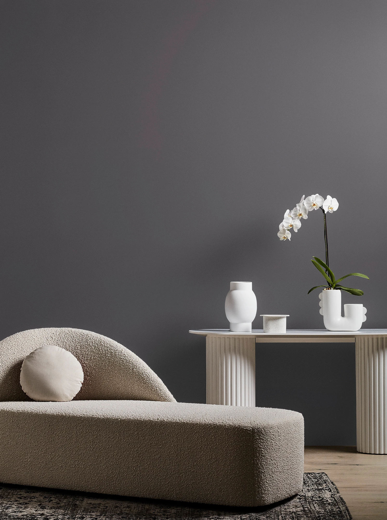 Neutral-coloured modern chaise longue and white table with orchid plant and vase in grey living room. 