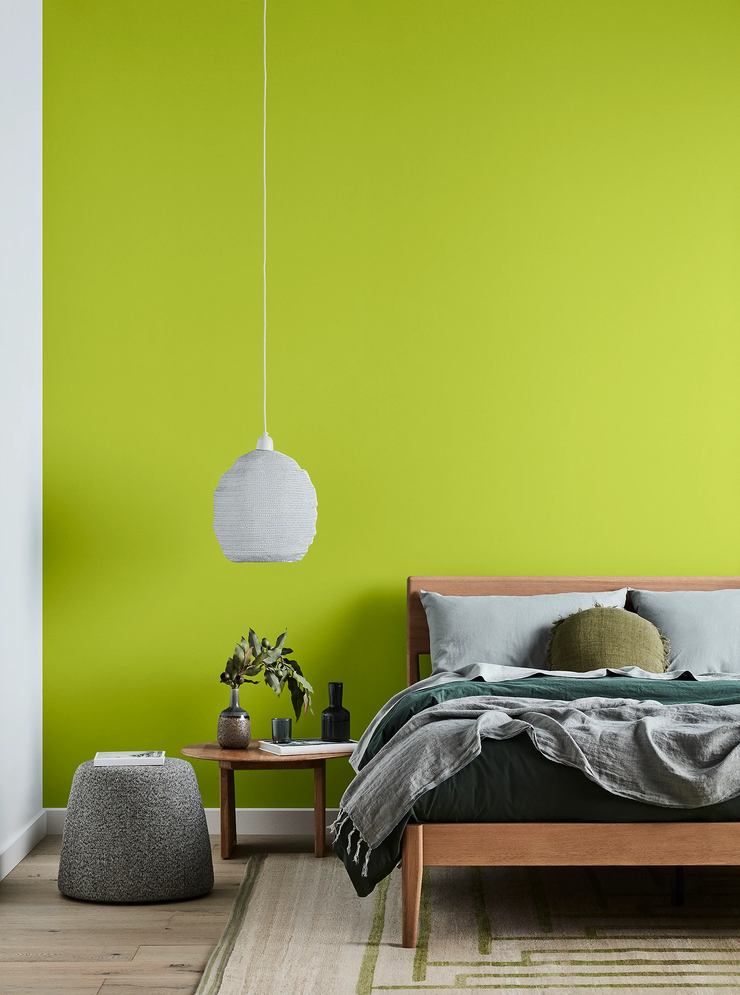 Green bedroom with timber bed and side table, white pendant light and grey linens.