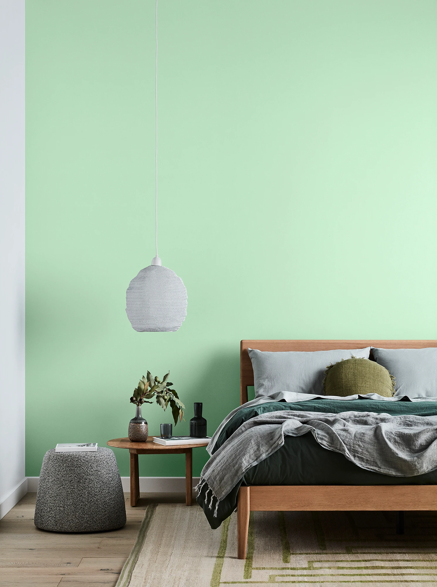 Green bedroom with timber bed and side table, white pendant light and grey linens.