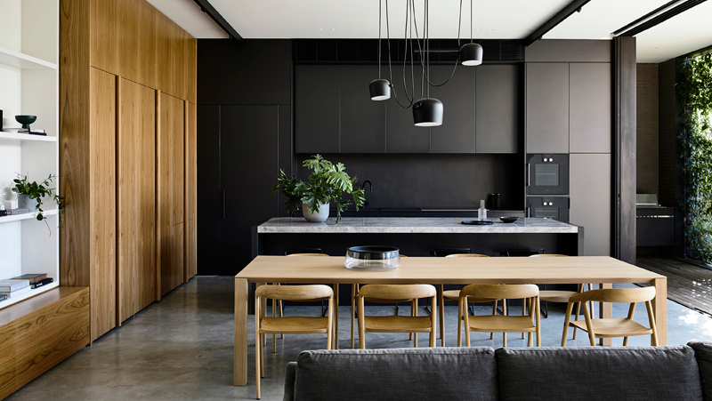 Open plan kitchen and dining area; black kitchen cabinets on end wall; timber kitchen cabinets on side wall; island bench; rectangle timber dining table with eight chairs.