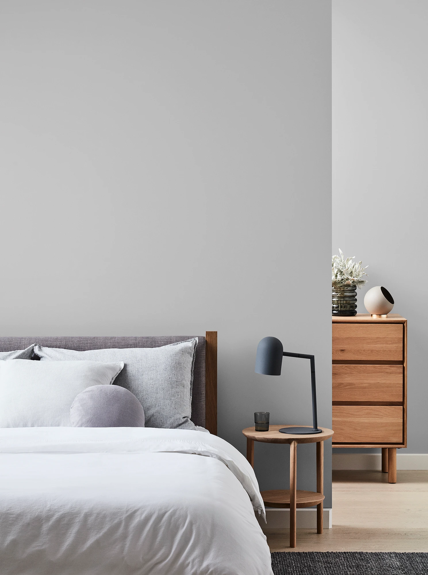 Grey bedroom with wooden chest of drawers and bedside table with black lamp and neutral coloured bed linen.