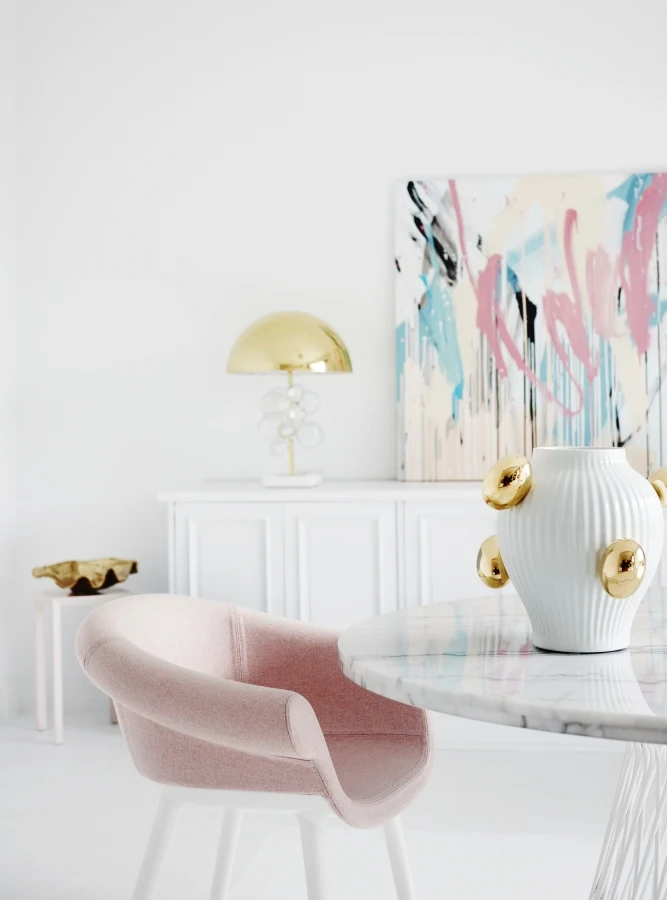 White dining room with glass and gold lamp on a white sideboard. Pink dining chair at white marble table with white and gold vase.