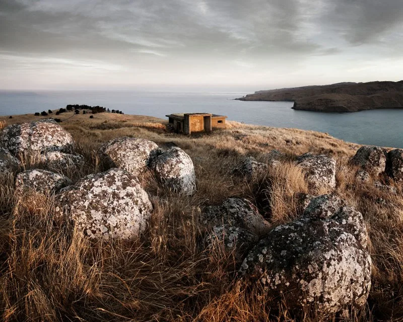 A wooden hut on a rocky mountain with large rocks and grasses overlooking water