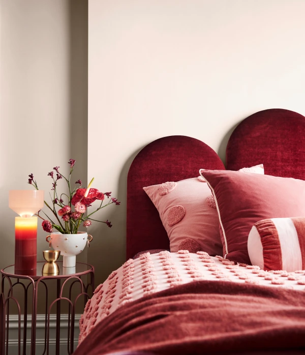A red and pink-toned decorated bed with a small glass side table featuring a candle and vase of flowers against a wall painted in Dulux White Dune Quarter.