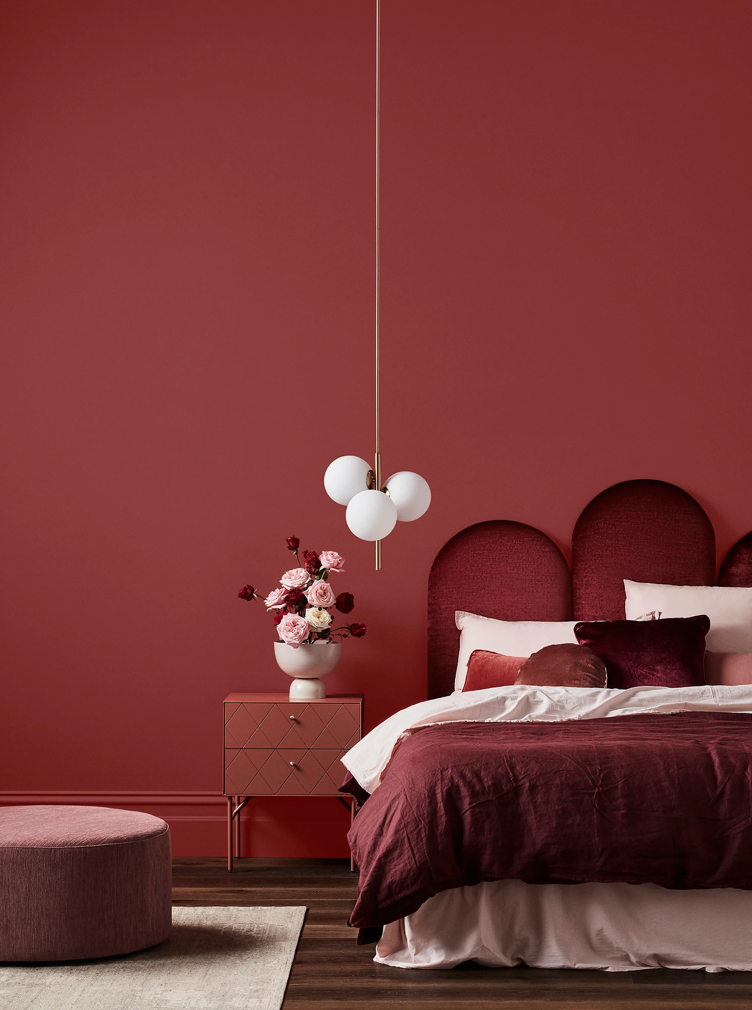 Deep red bedroom with maroon velvet headboard, pink bedside drawers, white pendant light and pink foot rest.