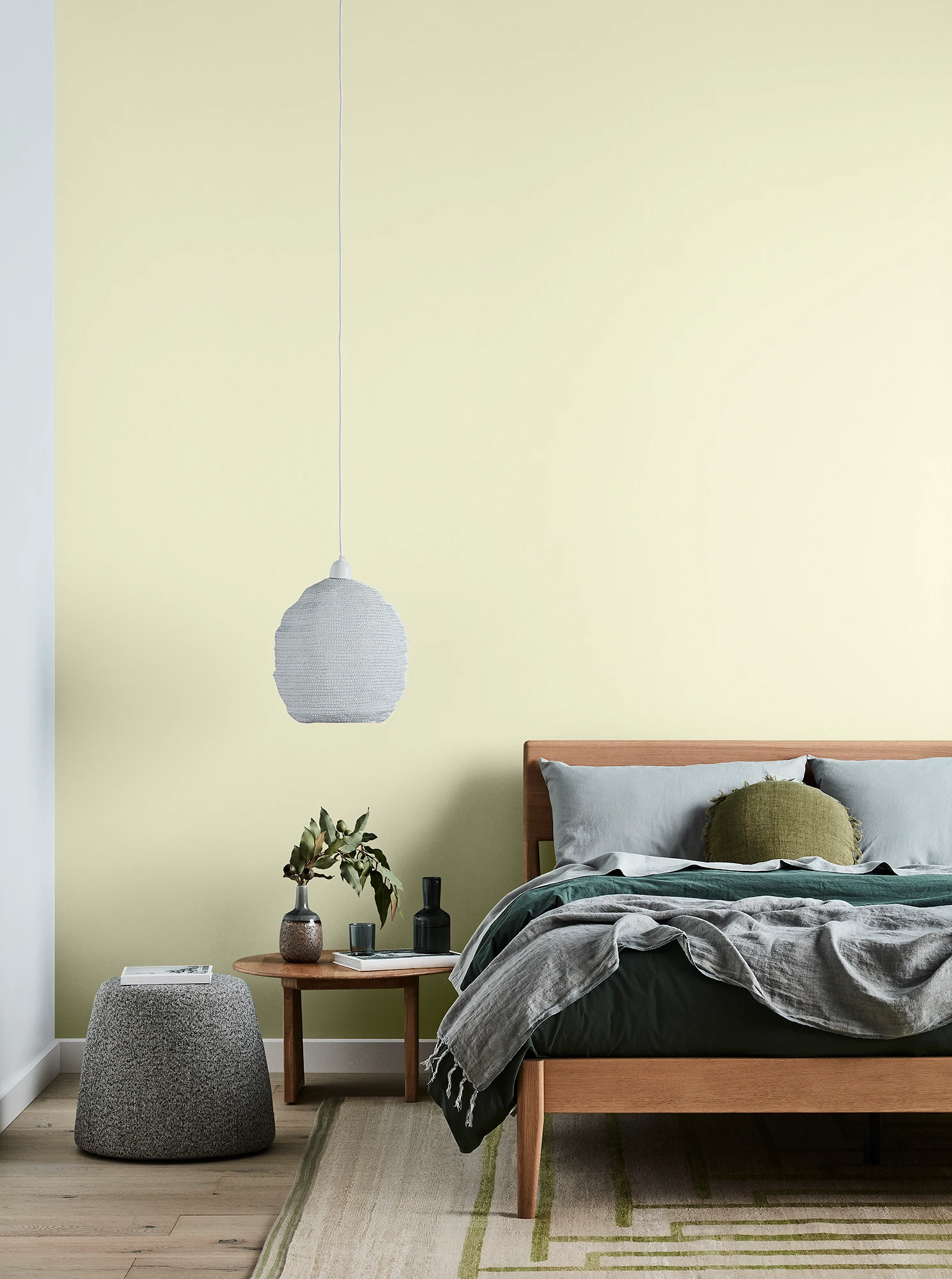 Green bedroom with timber bed and side table, white pendant light and grey linens.