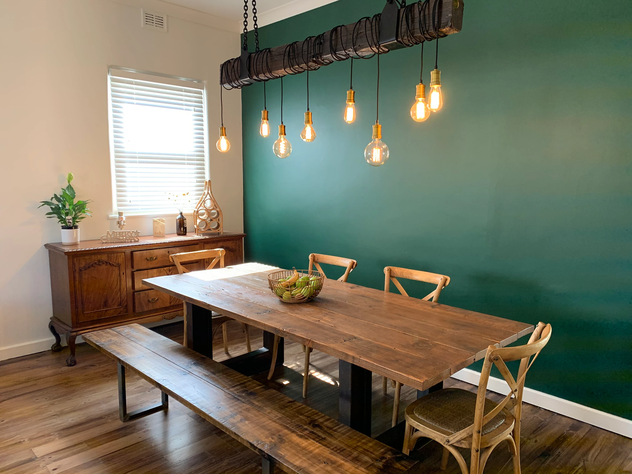 Dining room with dark green feature wall, rectangle timber table and light bulbs handing from suspended wooden beam