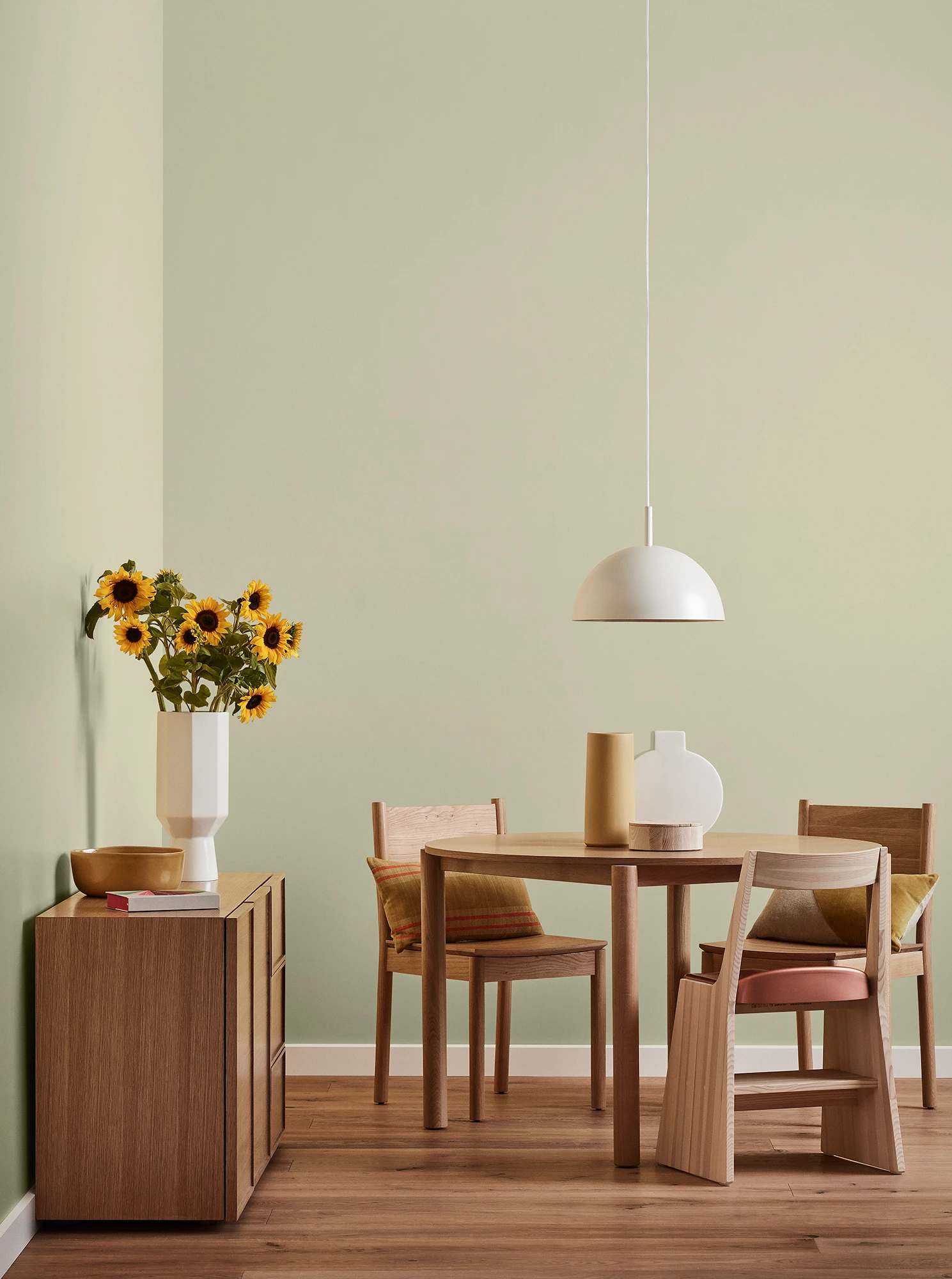 Round wooden table, three chairs and console with vase of sunflowers and white pendant light in neutral room.