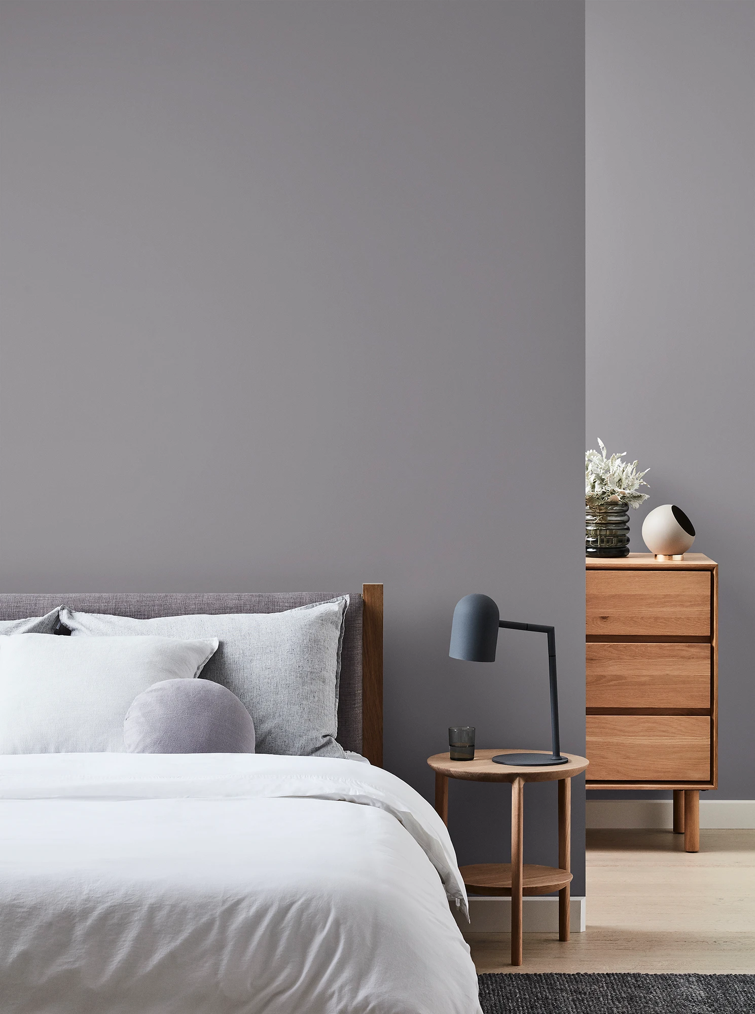 Grey bedroom with wooden chest of drawers and bedside table with black lamp and neutral coloured bed linen.