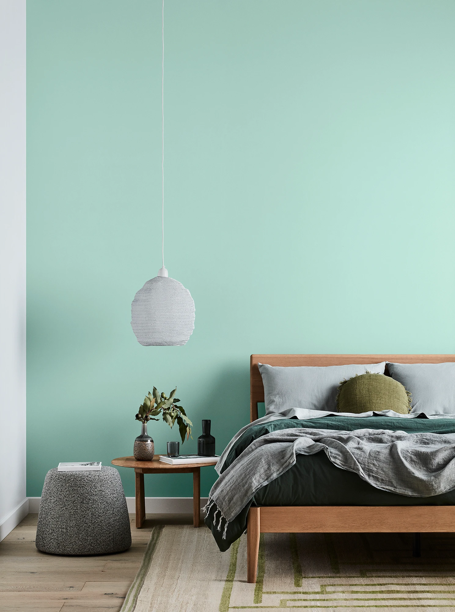 Green bedroom with timber bed and side table, white pendant light and grey linens.