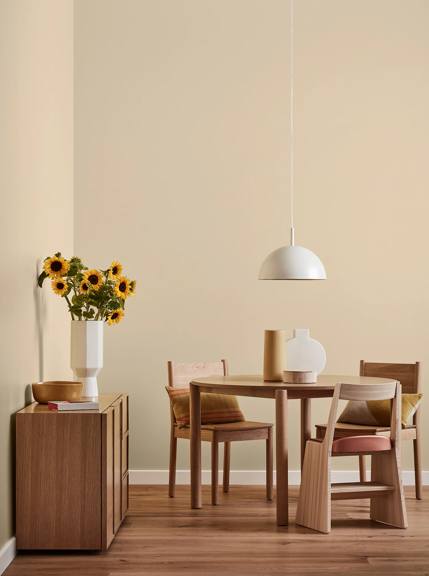 Round wooden table, three chairs and console with vase of sunflowers and white pendant light in neutral room.