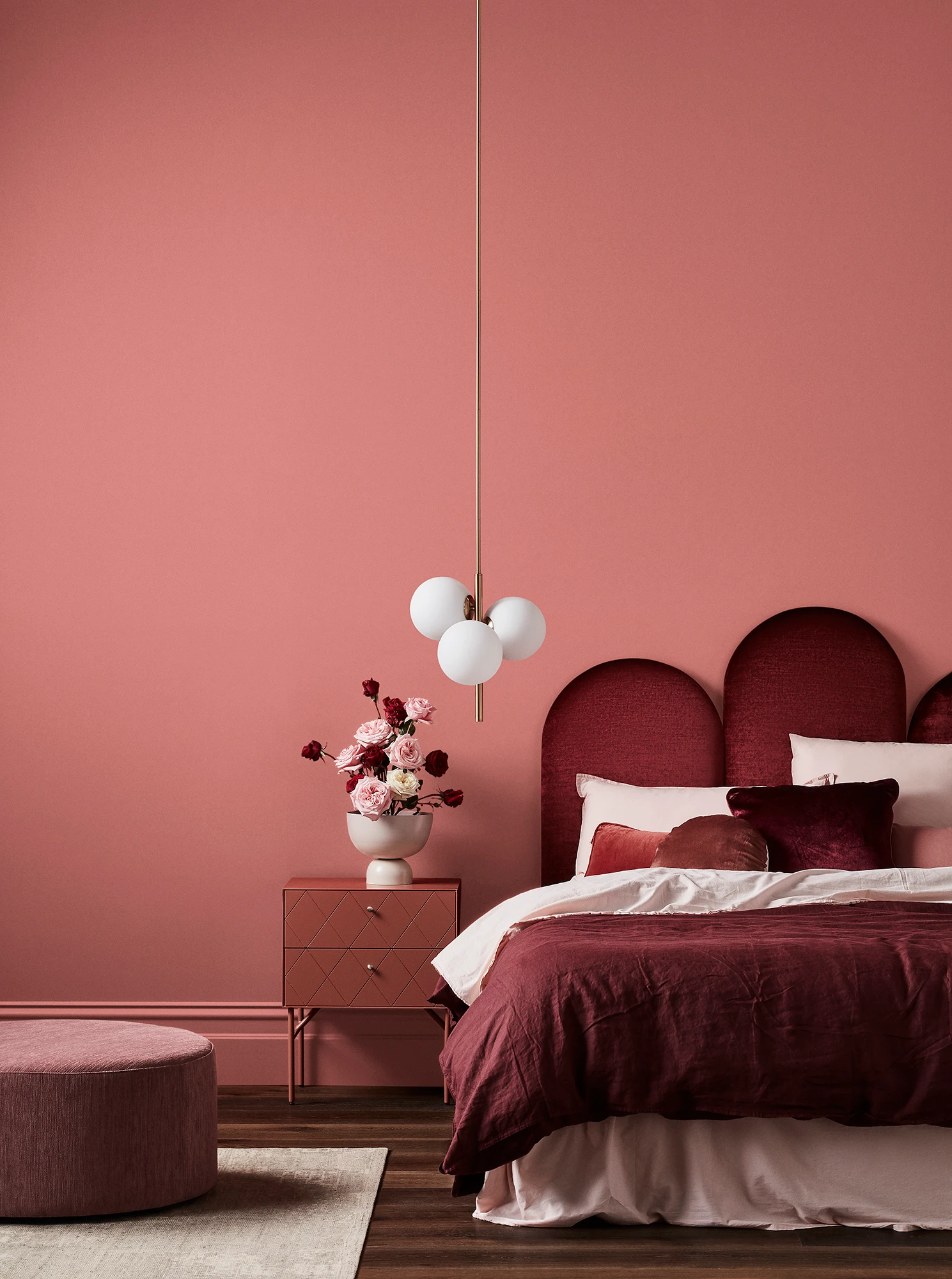 Pink bedroom with white pendant light, maroon velvet headboard, maroon and pink linens, timber bedside table and pink footrest.