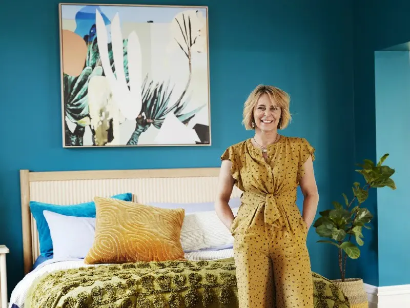 Woman standing in teal bedroom with timber bedhead and modern art on wall.