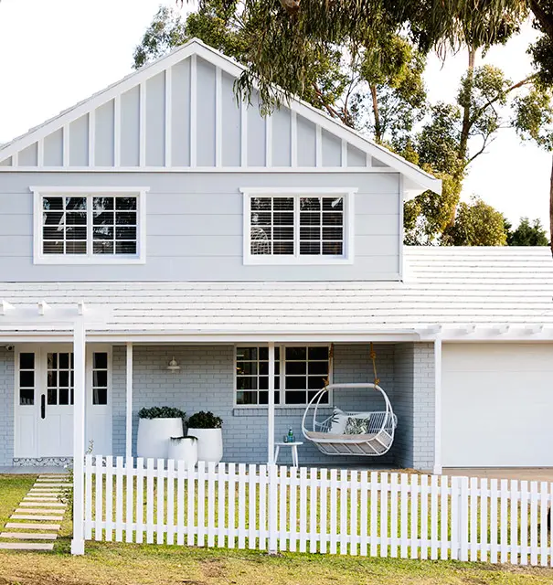 Hamptons style home with white picket fence out front and swinging chair on porch.