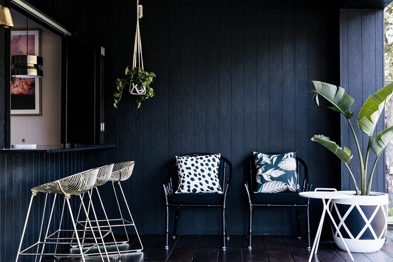 Black patio. Outdoor/indoor bar through window with three metal bar stools. Two black chairs with black and white cushions. Banana palm in large black and white pot, small round side table.