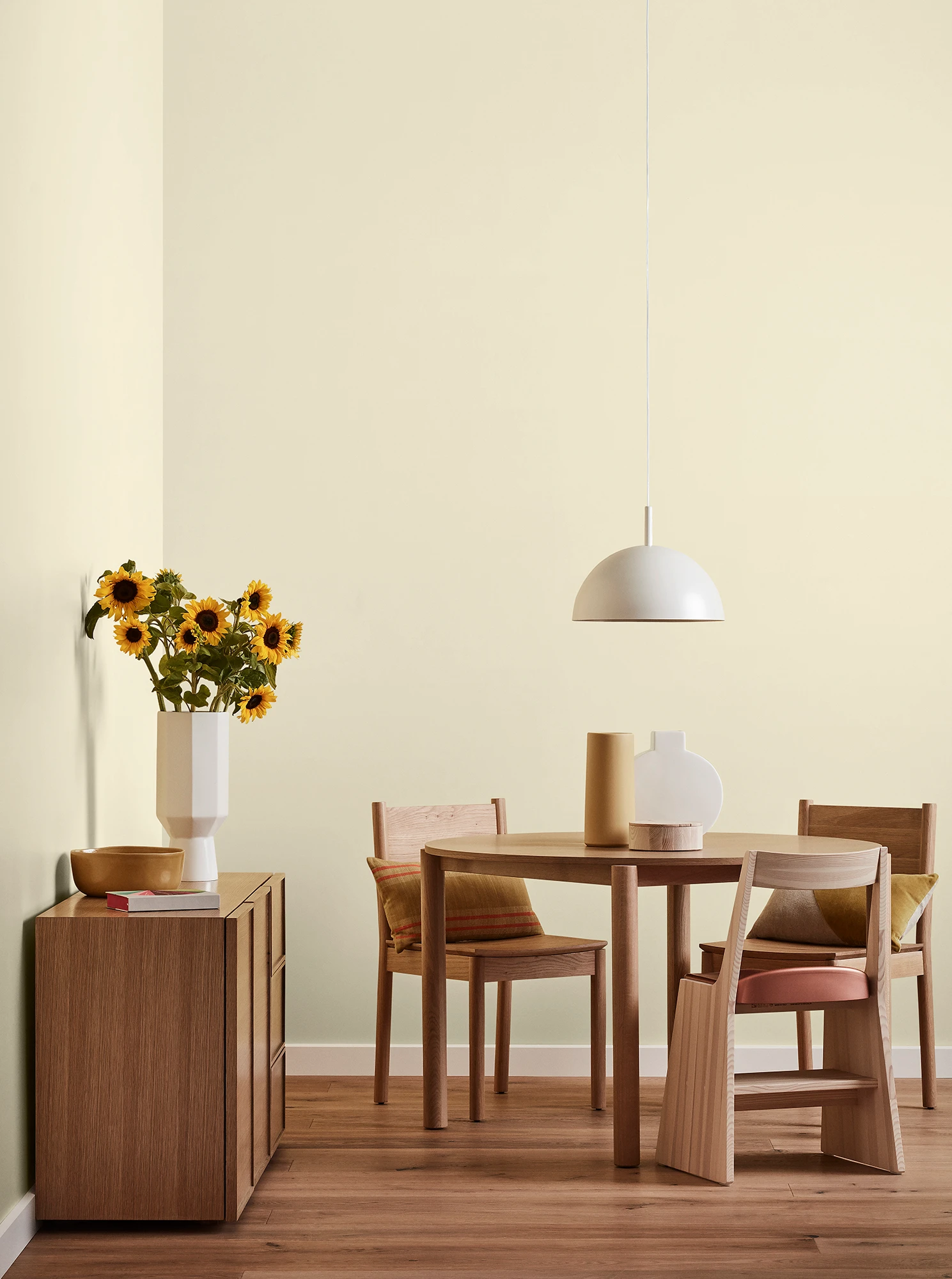 Round wooden table, three chairs and console with vase of sunflowers and white pendant light in neutral room.