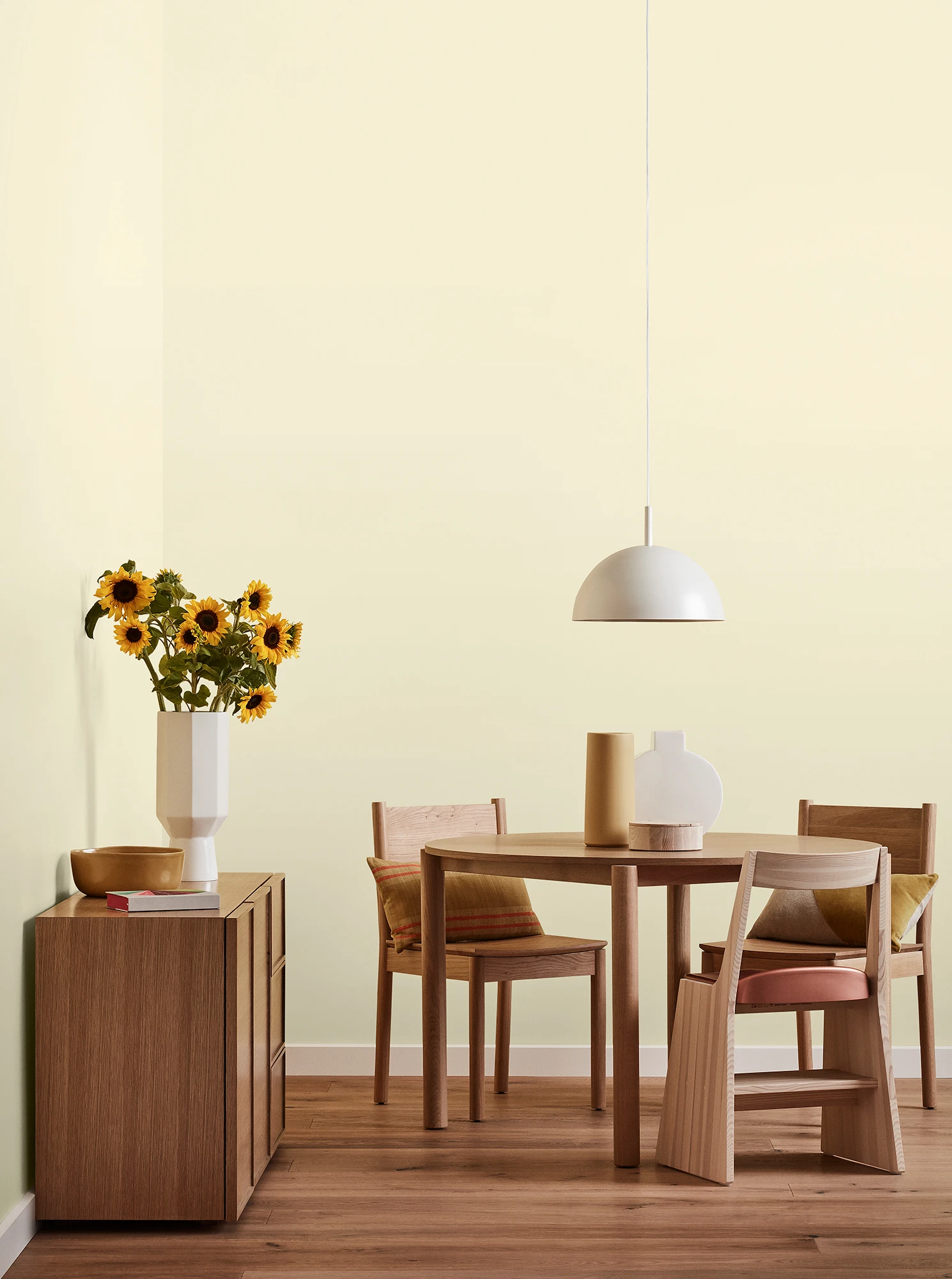 Round wooden table, three chairs and console with vase of sunflowers and white pendant light in light yellow room.