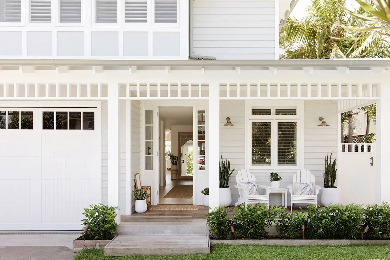White exterior with chairs on a front deck, a garage door and open front door looking into a hallway with a mirror at the end