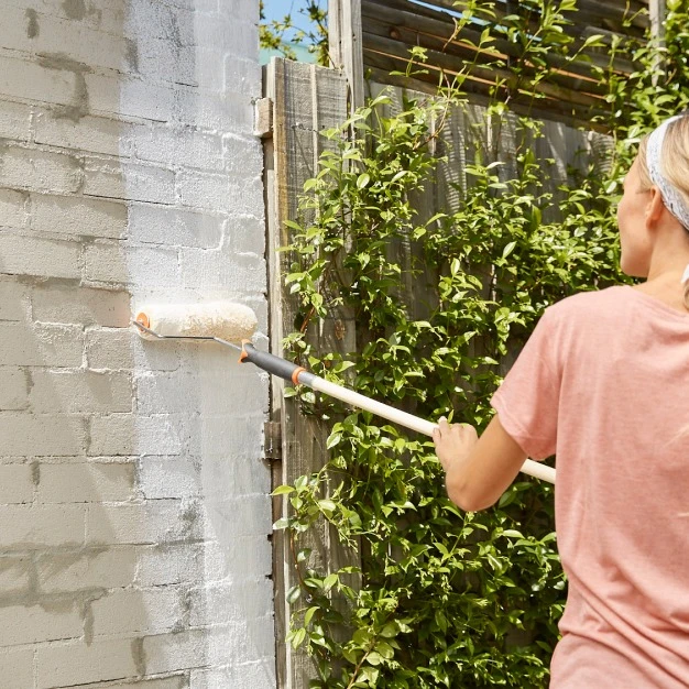 Woman painting brickwork outside in a t-shirt with green shrubbery behind her.