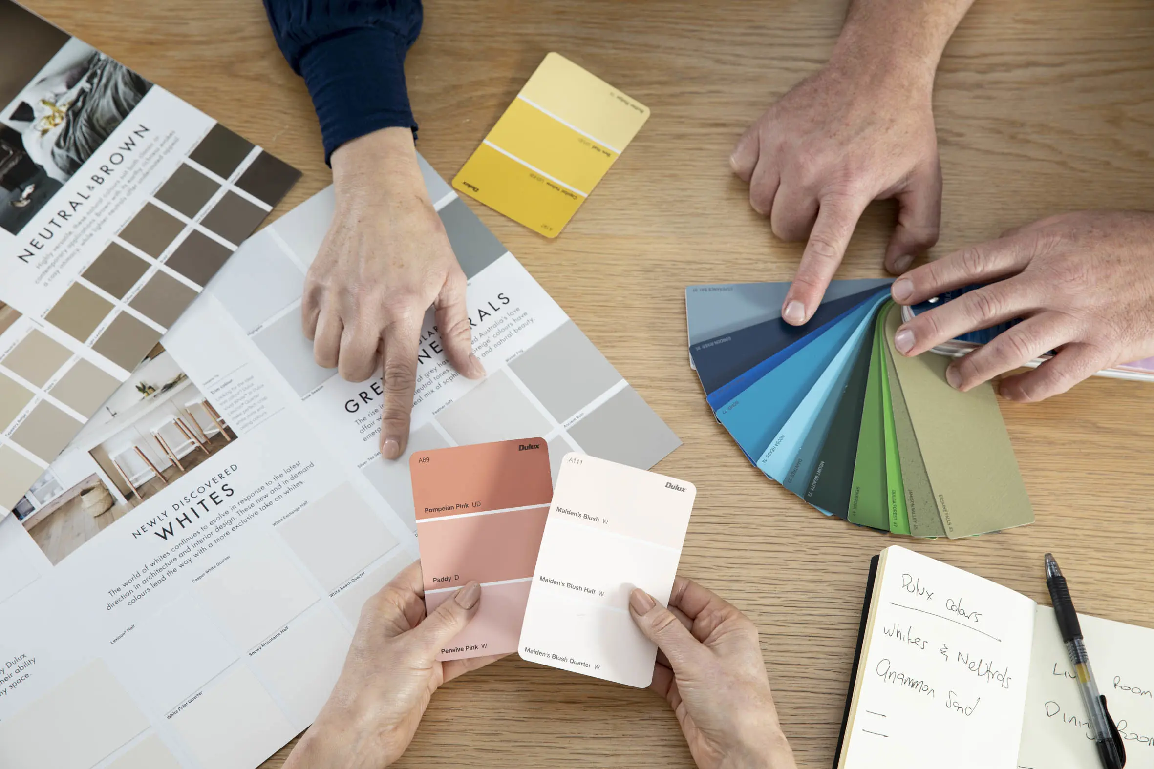 Three hands holing Dulux colour chip cards displaying orange, whites and yellow schemes. 