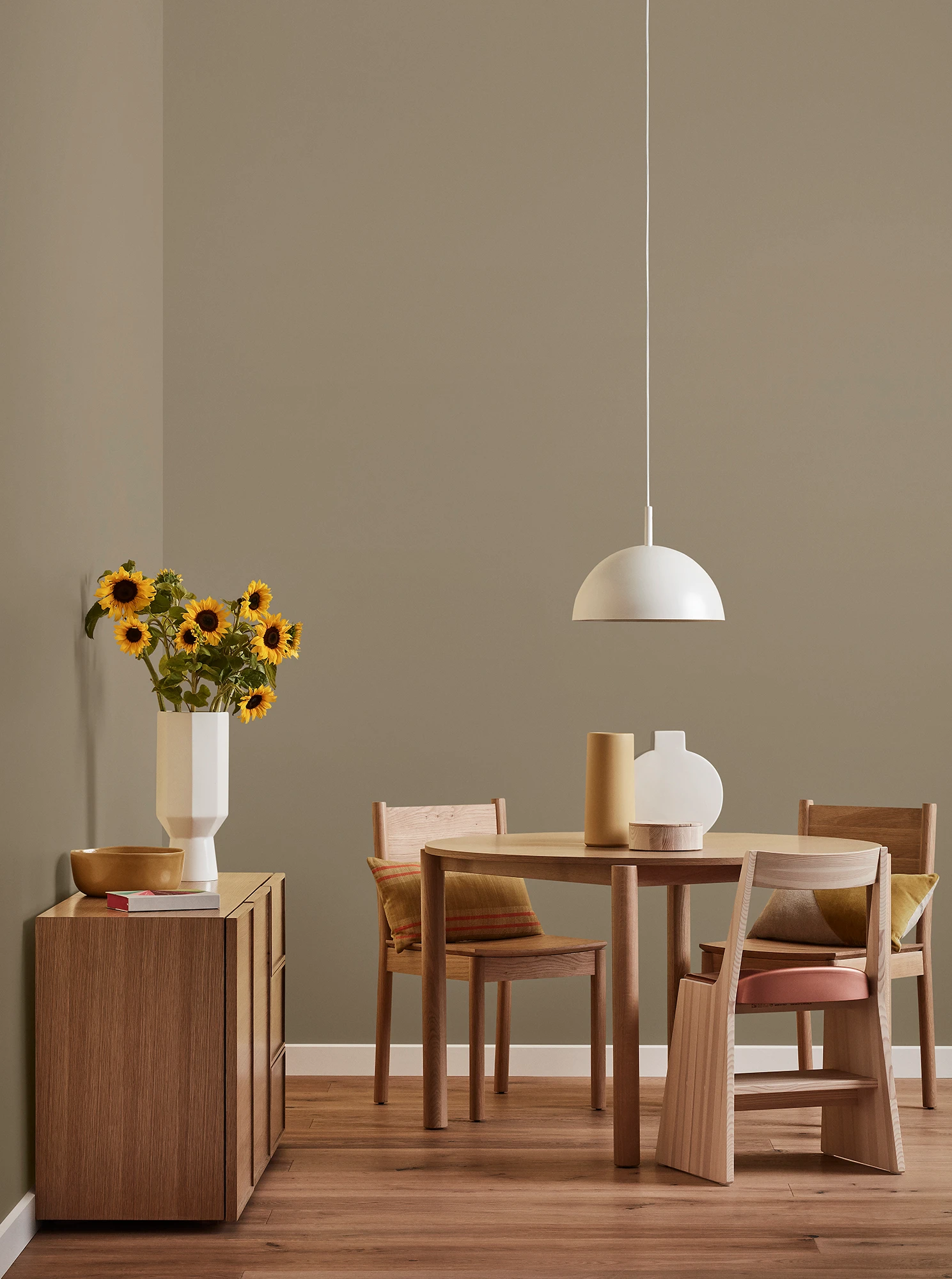 Round wooden table, three chairs and console with vase of sunflowers and white pendant light in neutral-brown room.