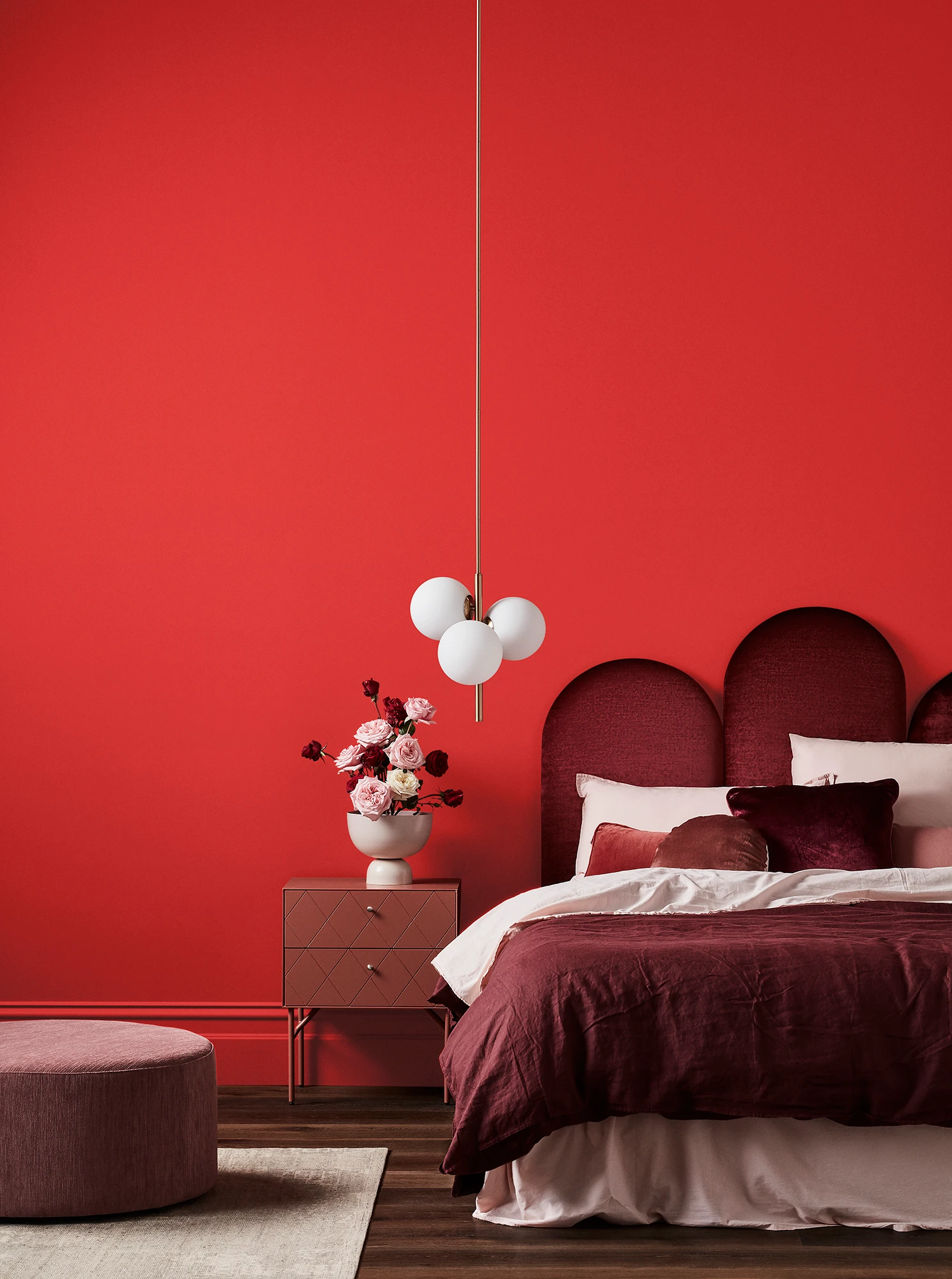 Red bedroom with maroon velvet headboard, pink and maroon linens, white pendant light and timber bedside drawers