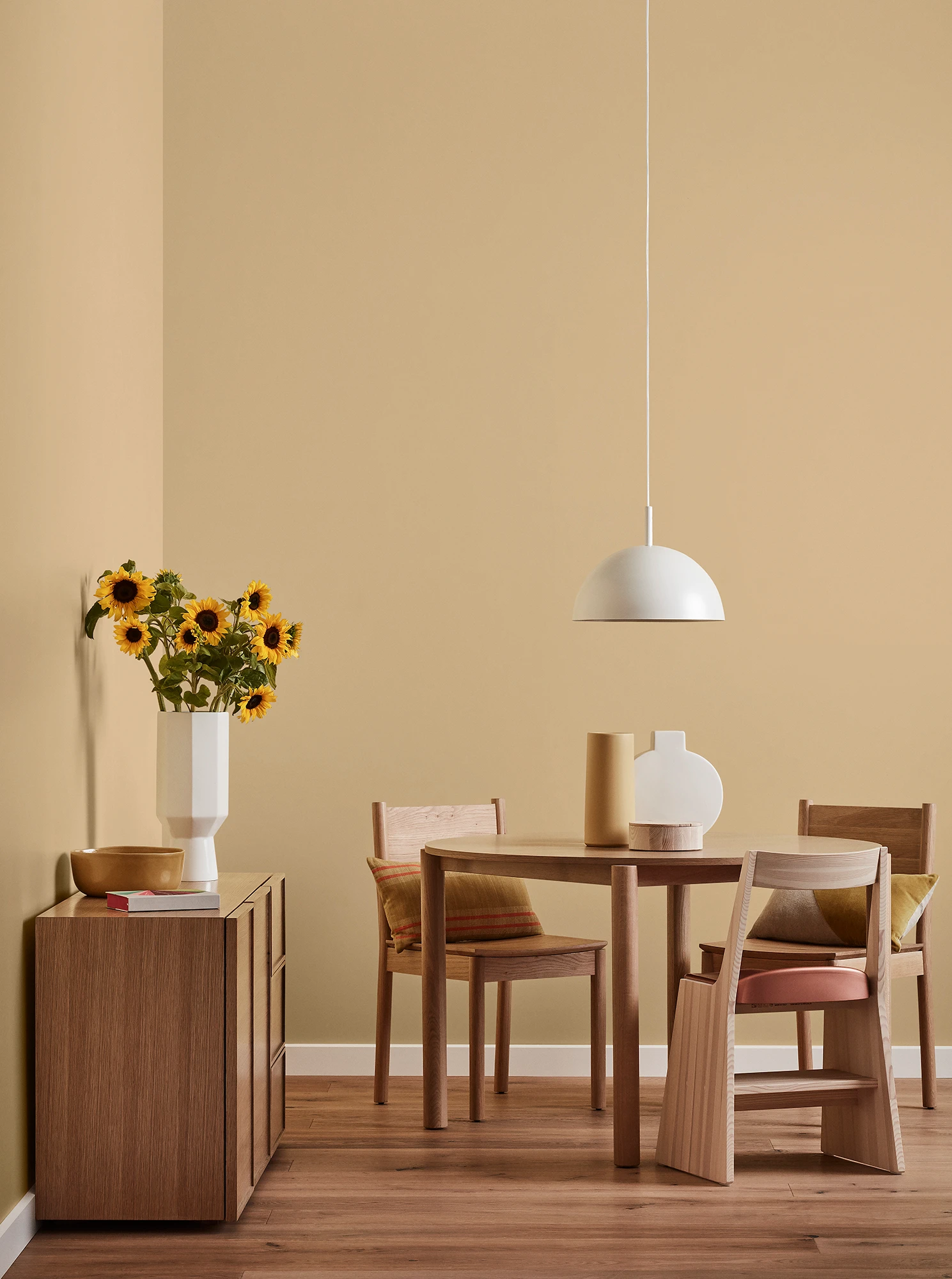 Round wooden table, three chairs and console with vase of sunflowers and white pendant light in neutral room.