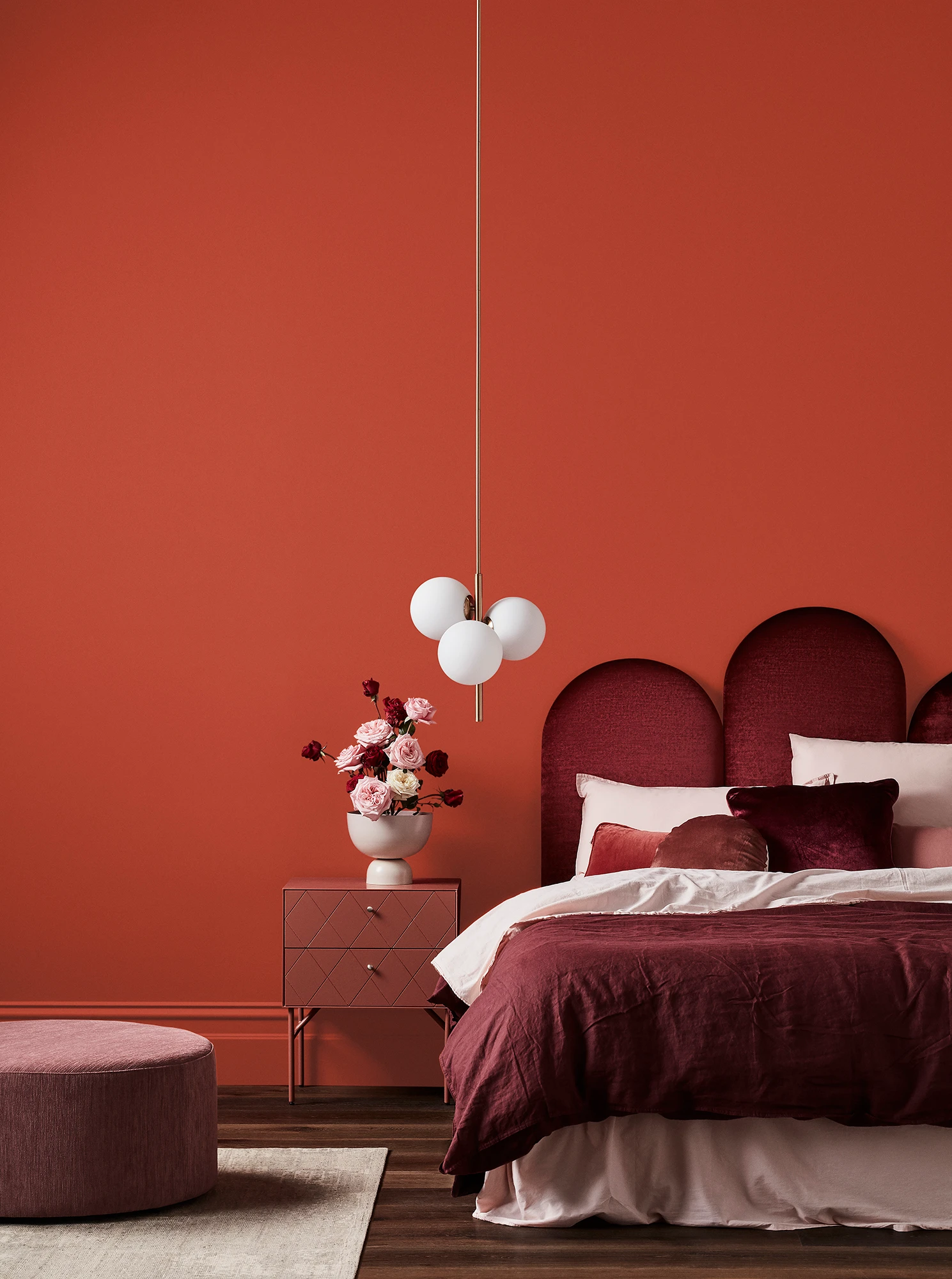 Orange-red bedroom with white pendant light, maroon velvet headboard, maroon and pink linens, timber bedside table and pink footrest.