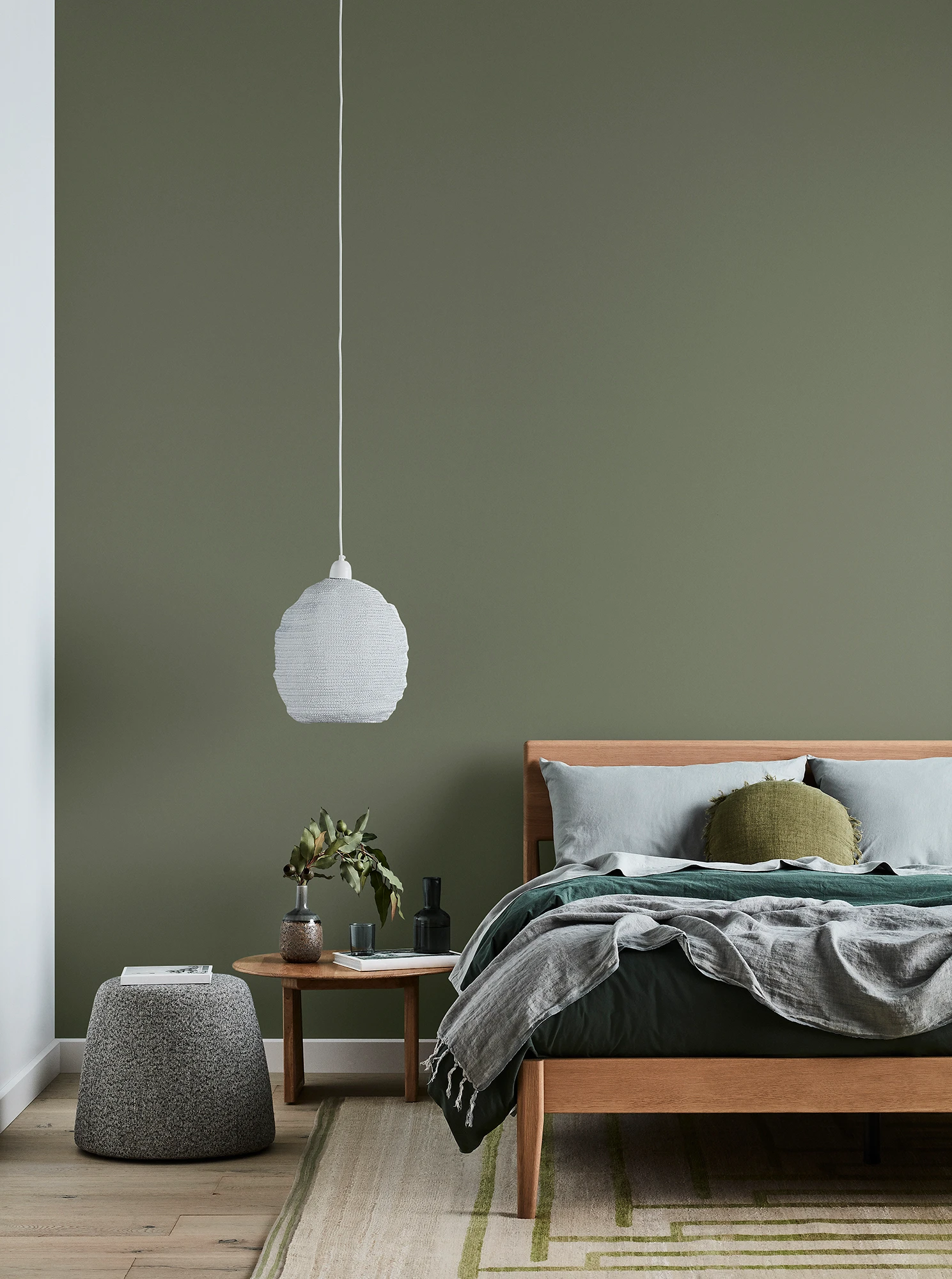 Green bedroom with timber bed and side table, white pendant light and grey linens.