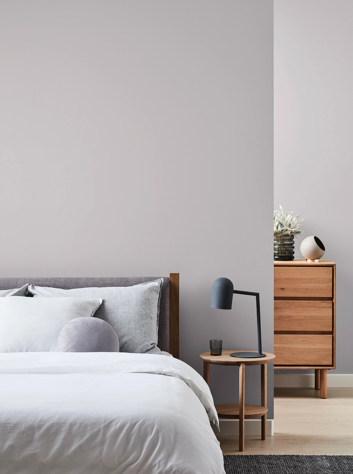 Grey bedroom with wooden chest of drawers and bedside table with black lamp and neutral coloured bed linen.