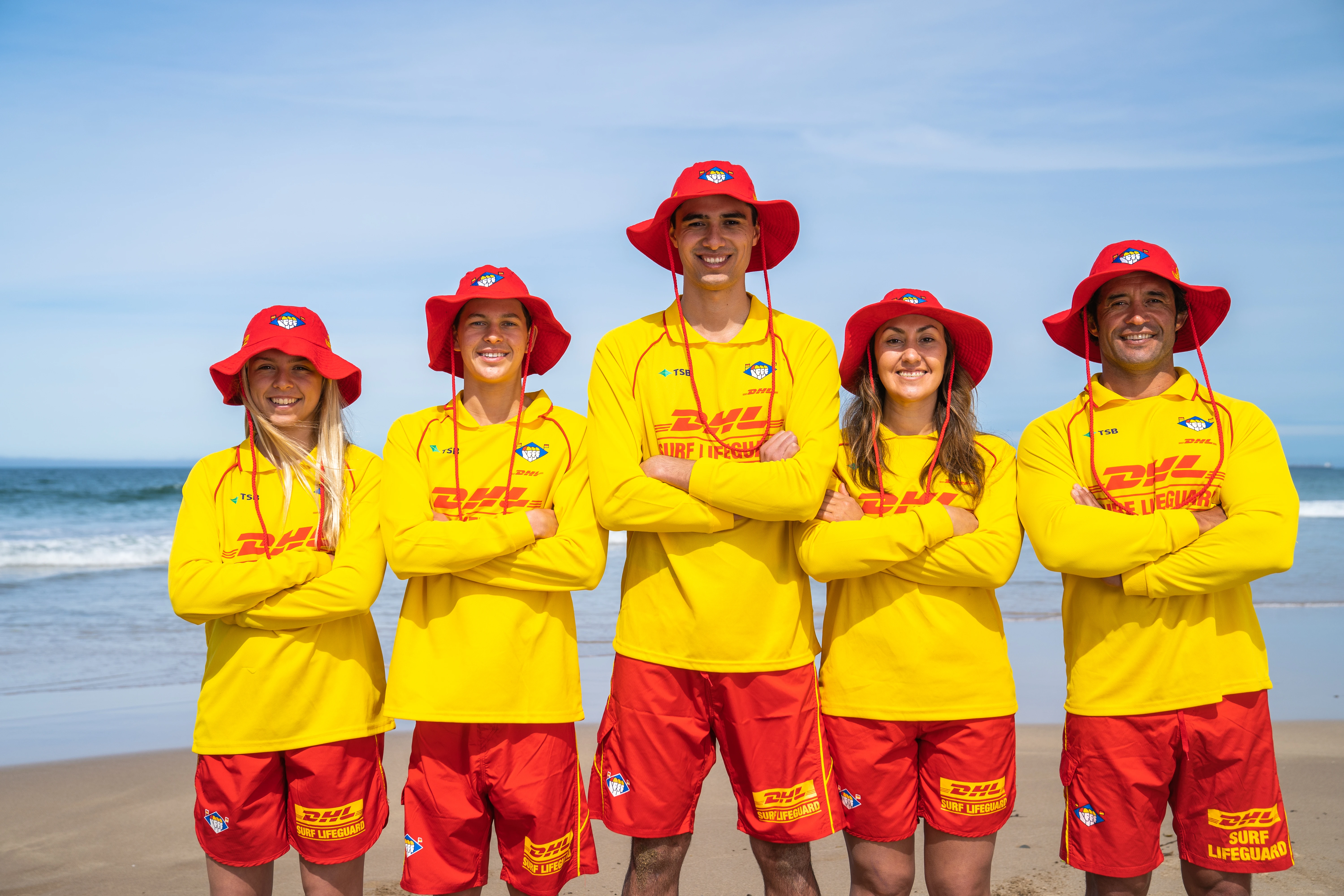 Five people standing in their surf life saving uniforms on a beach with their arms crossed