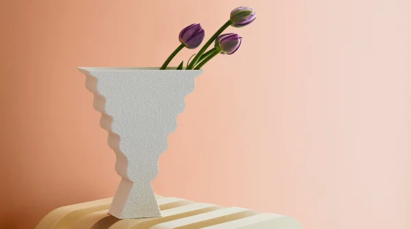 White inverted triangular vase with three pink tulips on yellow table with orange wall in background.