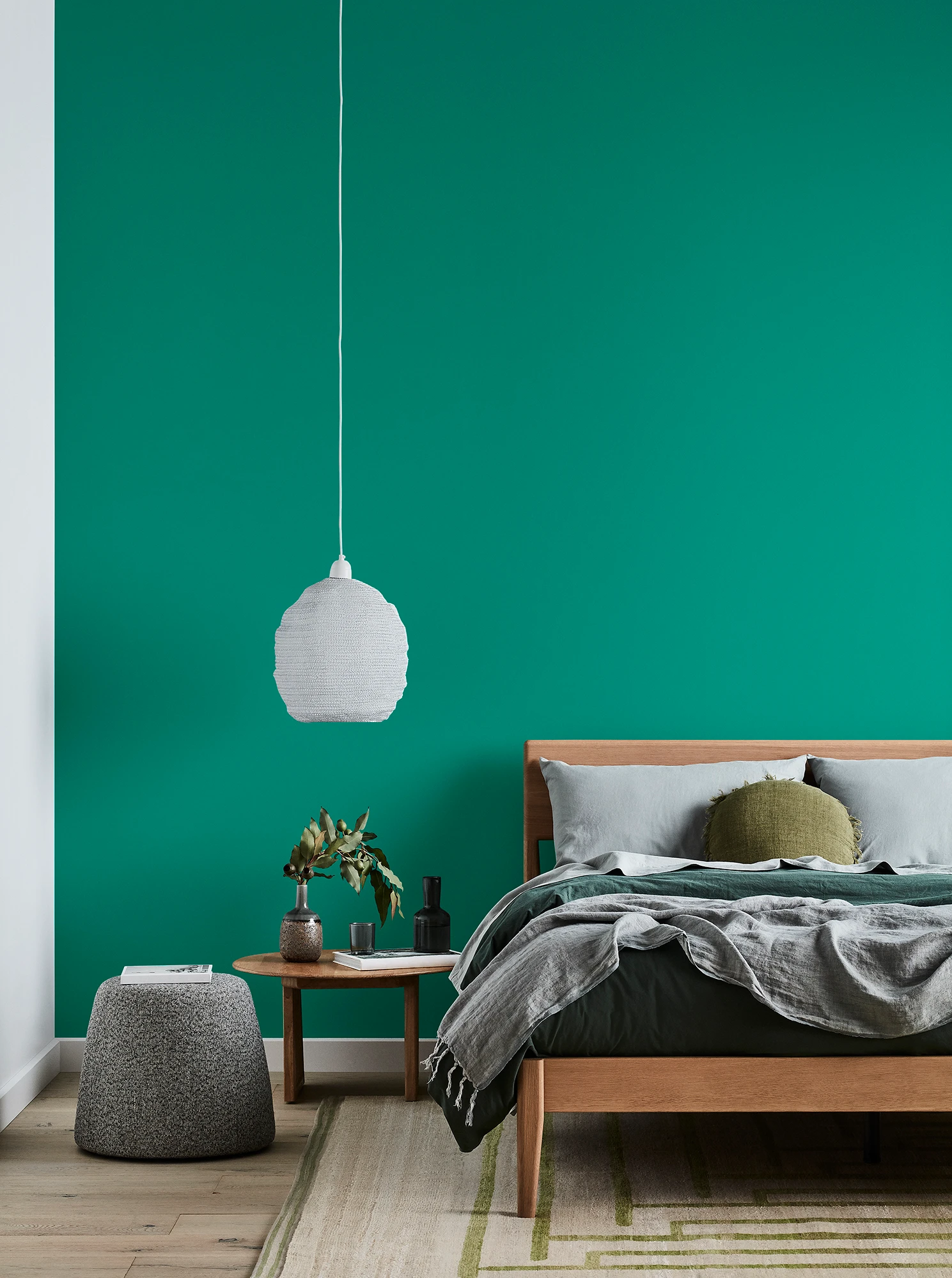 Green bedroom with timber bed and side table, white pendant light and grey linens.