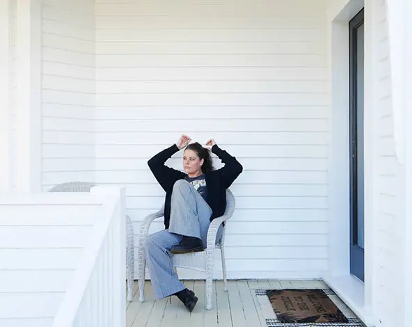 Jess sits on a chair outside the front of a weatherboard house painted in white