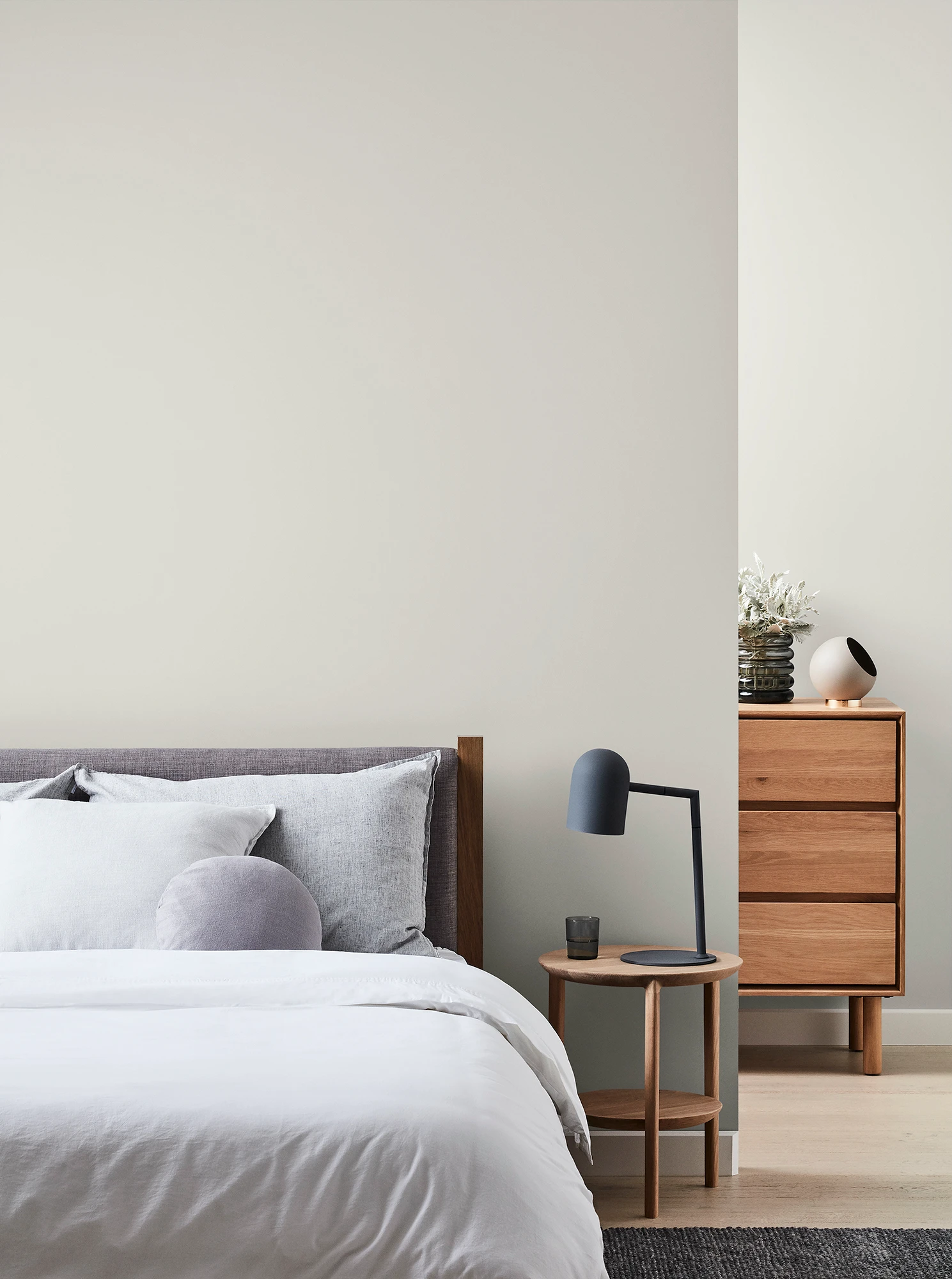 Grey bedroom with wooden chest of drawers and bedside table with black lamp and neutral coloured bed linen.