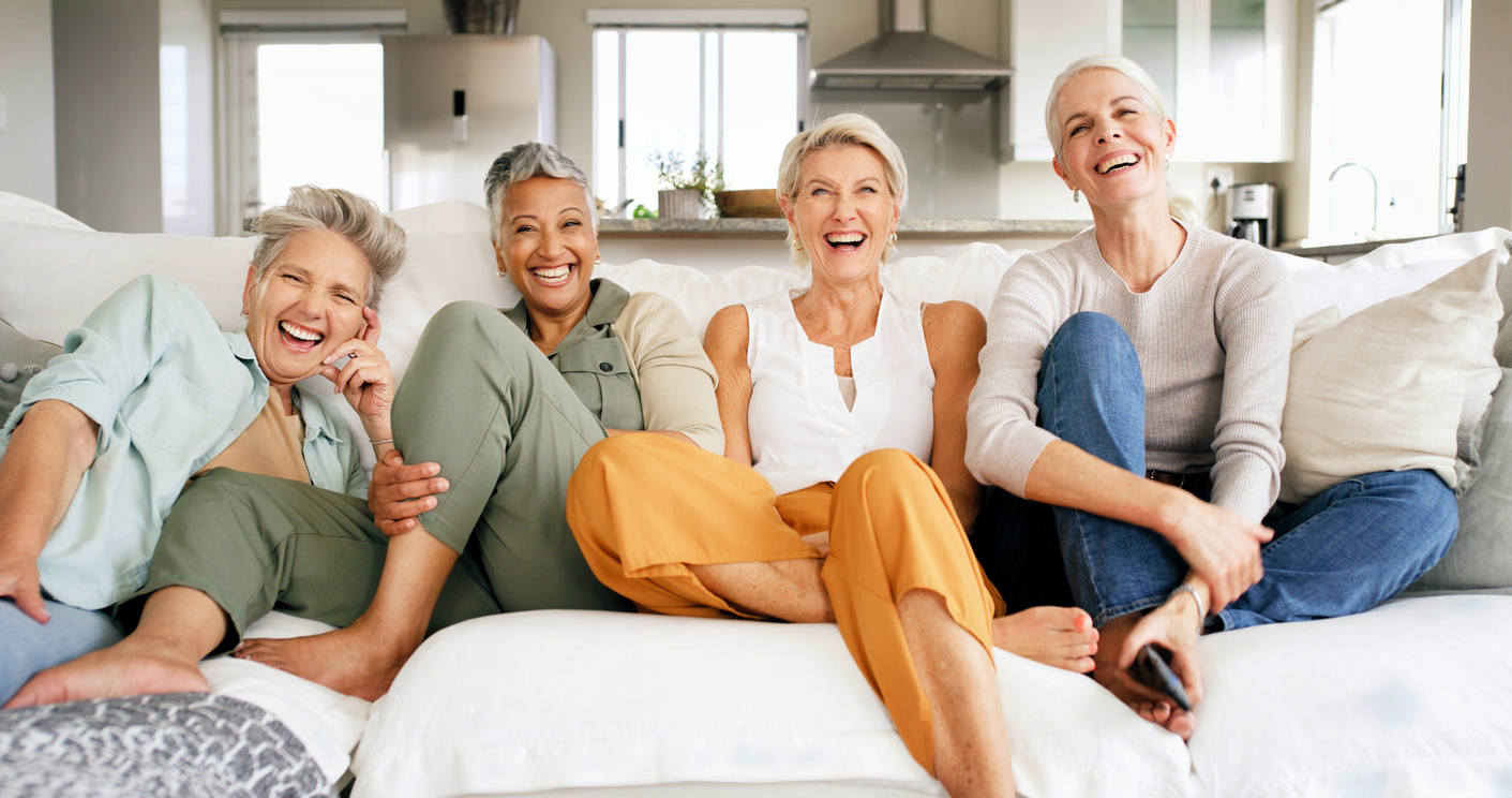 A group of older women sit together on a couch and smile into the camera