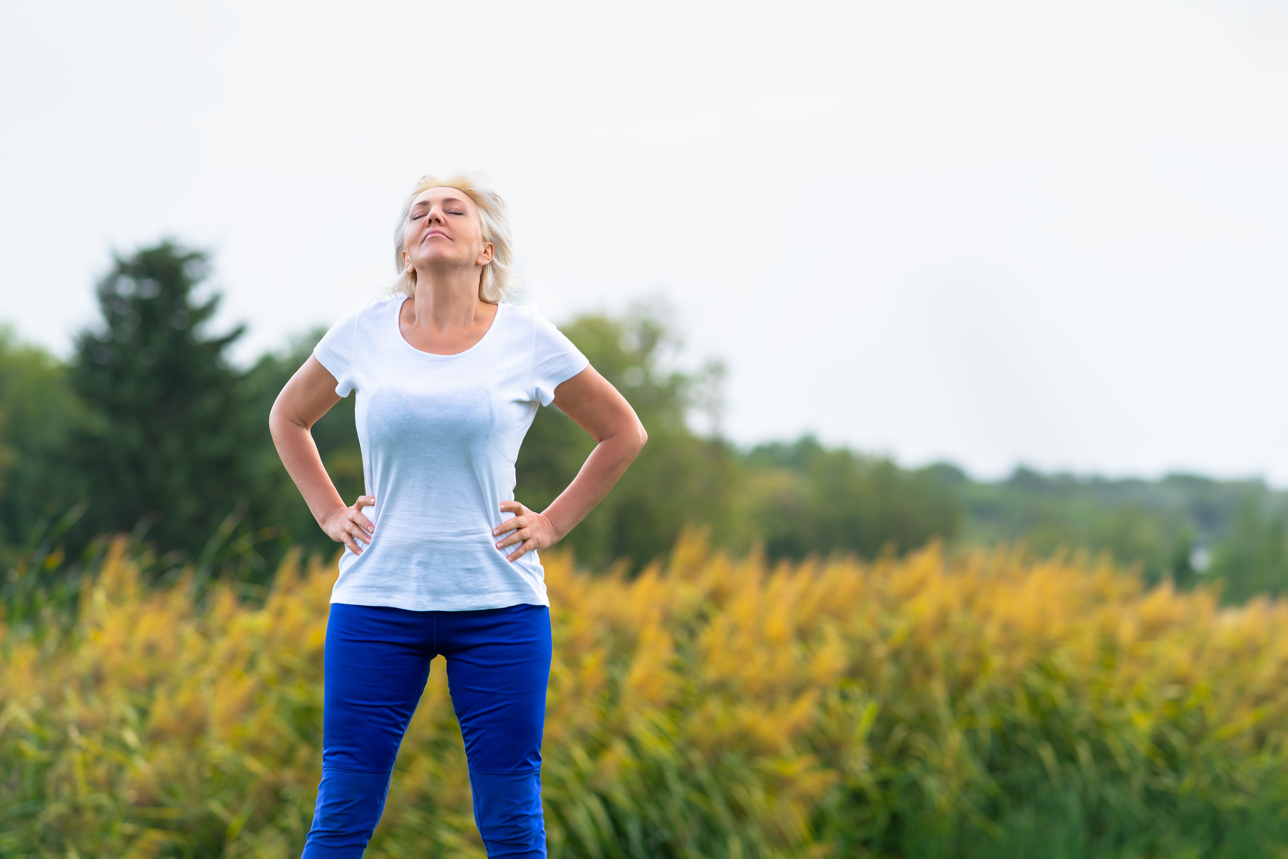 A woman with her hands on her hips closes her eyes and looks into the sky while standing in a field