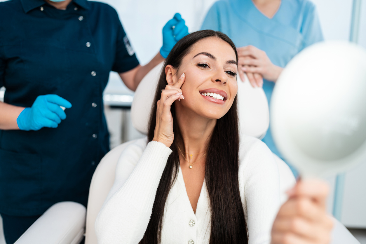 A woman in a provider office holds a mirror up to her face and touches her check