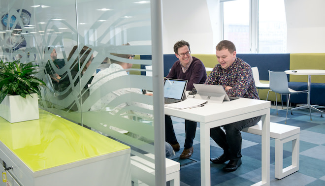 A picture of a communal area in an office. Facing the camera are two employees seating at a long table and bench. These two employees are chatting, smiling and looking into their laptop screens. You can also see other employees opposite those facing the camera, but they have their backs to the camera and are behind a panel of obscured glass. Other tables and chairs, office drawers, and pot plants can be seen in and near the communal office area.