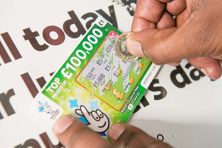 An individual that is holding a National Lottery scratchcard in one hand and a coin in the other. You can only see the individual's hands, the scratchcard and coin. The scratchcard is entitled 'Top prizes of £100,000' and has the National Lottery crossed fingers with eyes logo on the right. The individual is scratching off the scratchcard to reveal a number of monetary values.