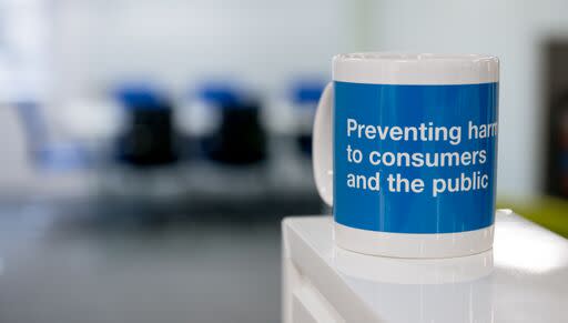 A picture of a mug in the foreground with the writing 'Preventing harm to consumers and the public'. The mug is predominantly blue with a white boarder and handle. In the background is a blurred meeting room with chairs in an office.